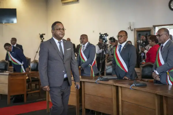 Des hommes en costume, certains portant des écharpes aux couleurs nationales, se tiennent dans une salle. L'un d'eux semble être une persona non grata, marchant à l'écart.